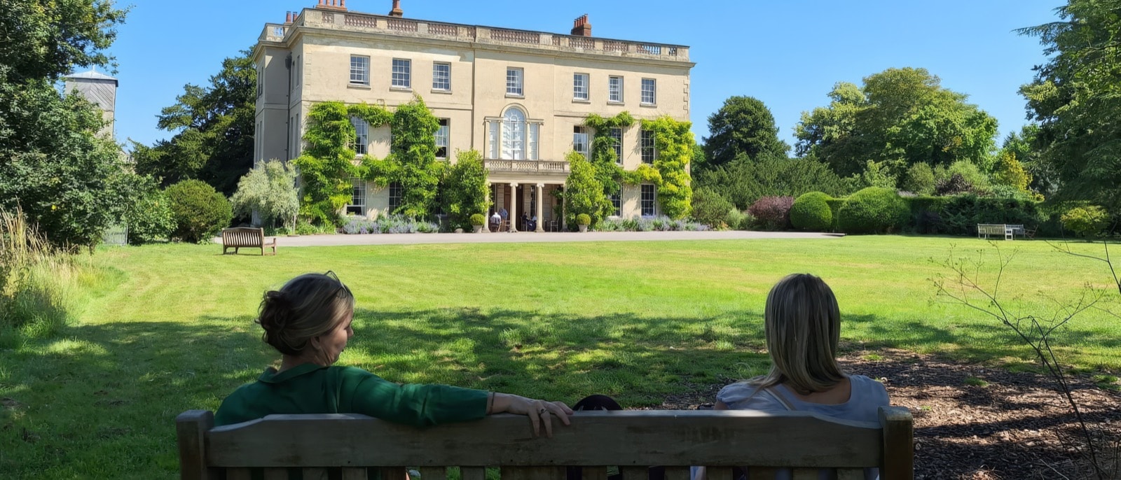 Two people sitting on a wooden bench looking across the lawn toward Waterperry House