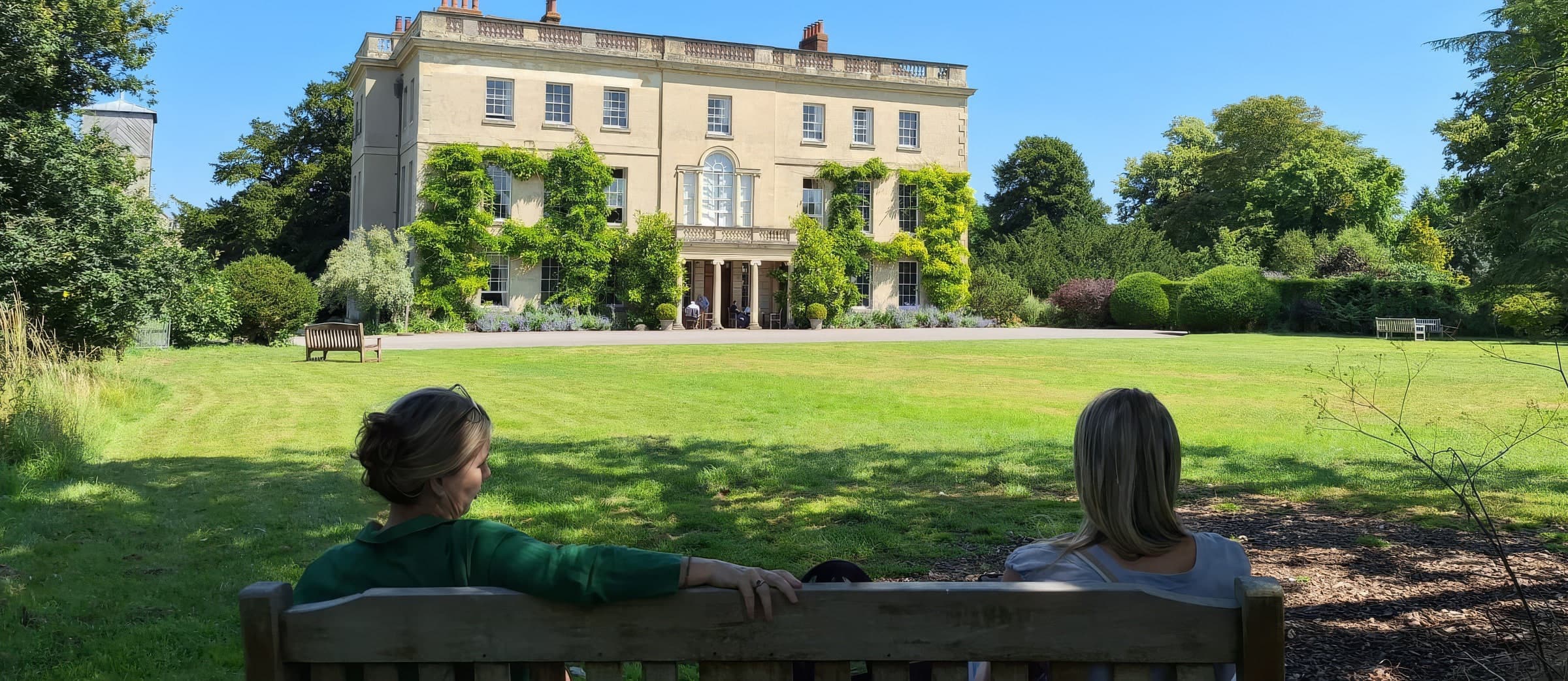 Two people seated on a garden bench looking across a sunlit lawn toward Waterperry House