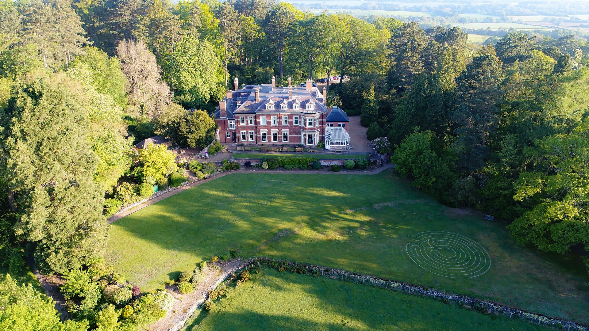 Aerial view of Nanpantan Hall and woodland with a circular grass labyrinth on the sloping front lawn