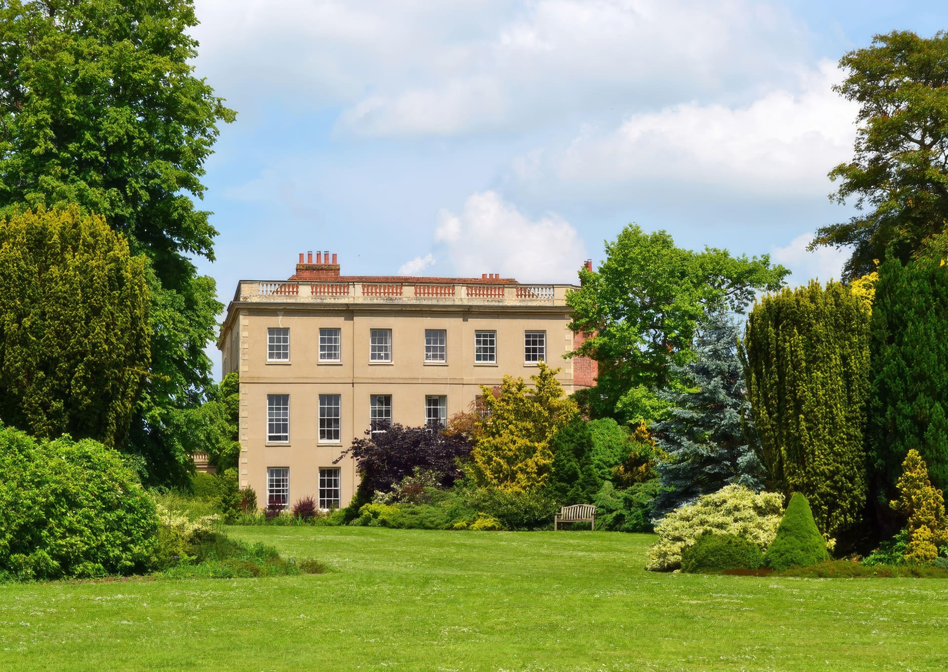 Waterperry House seen across a manicured lawn, framed by mature trees and garden planting under a blue sky