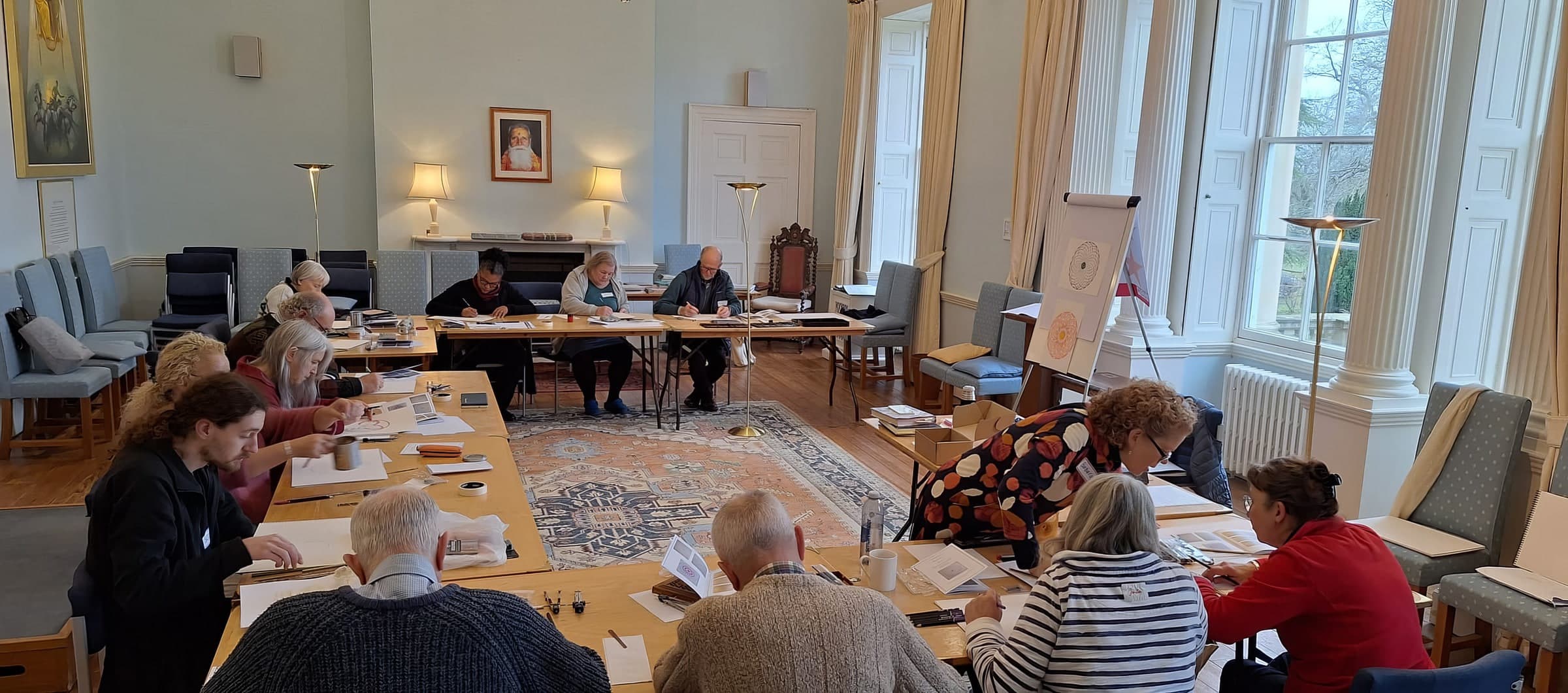 Participants drawing geometric patterns with compass and straightedge at long tables in a sunlit hall